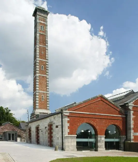 A historic red-brick building with arched windows and a tall, narrow chimney stands under a blue sky with scattered clouds. Trees and smaller buildings are visible in the background.