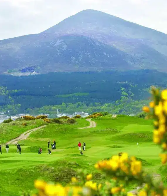Golfers play on a lush green course surrounded by yellow flowering bushes, with a scenic backdrop of forested hills and a tall, misty mountain under a cloudy sky.