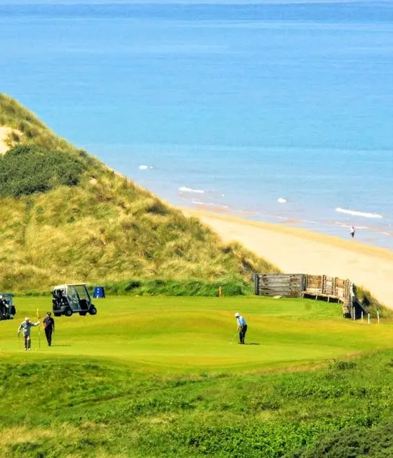 Golfers play on a green nestled between grassy dunes near the sea, with golf buggies nearby and a sandy beach stretching along the blue water in the background.