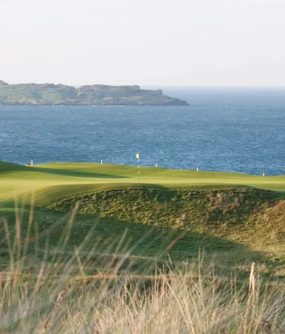A coastal golf course with rolling green hills in the foreground, a flag marking a hole, and the sea with an island in the distance under a clear sky.