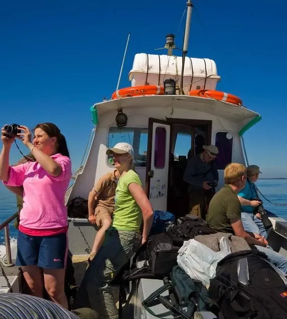 A group of people sit and stand on a small boat under a clear blue sky, some looking at the scenery while one person in a pink shirt takes a photo. Several bags and rucksacks are on the deck.