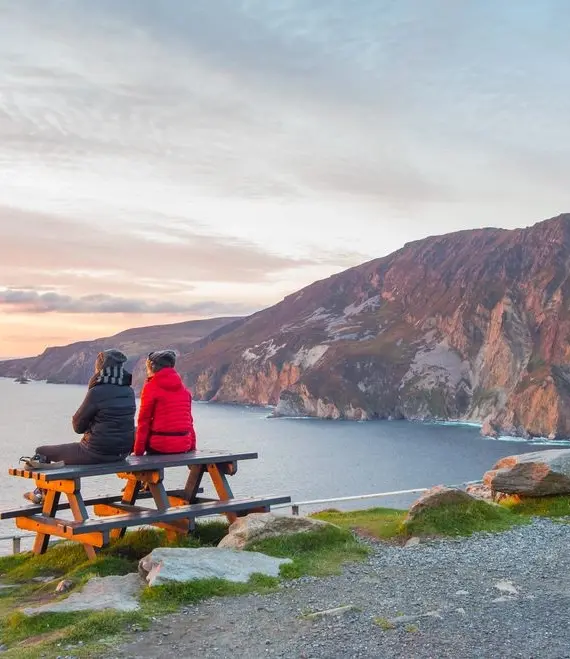 Two people wearing jackets sit on a picnic table overlooking cliffs and the sea at sunset, with dramatic skies and rugged coastline in the background.