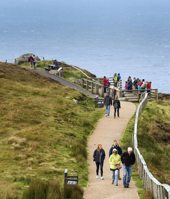 A group of people walk along a paved path with railings on a grassy cliffside overlooking the sea under an overcast sky. Some stop to view the scenery at a vantage point.