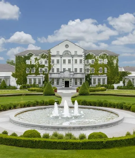 A large, elegant white building covered partly in ivy sits behind a circular fountain with three water jets, surrounded by manicured lawns, hedges, and landscaping under a blue sky with scattered clouds.