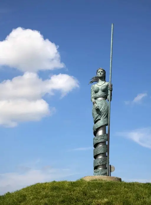 A bronze statue of a woman holding a tall spear stands on a grassy hill under a blue sky with white clouds. The figure's dress appears to spiral around her legs.
