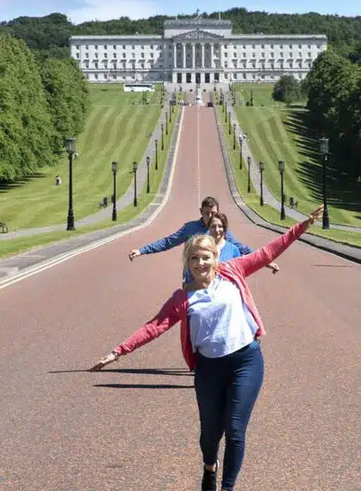 Three people stand in a row with arms outstretched on a wide path leading to a large white building with columns, surrounded by green lawns and trees on a sunny day.