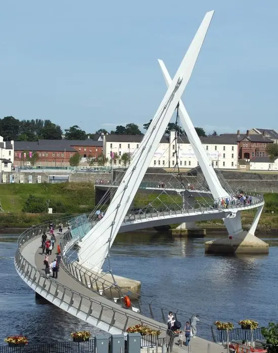 A modern, curved pedestrian bridge with two tall, angled white supports spans a river. People are walking across the bridge, and buildings and trees are visible in the background.