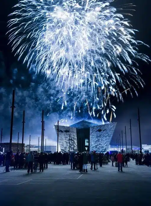 A large crowd gathers at night in front of a modern, angular building as bright white fireworks explode in the sky above, illuminating the scene.