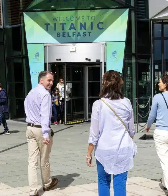 Three adults walk towards the entrance of Titanic Belfast, whilst other people walk nearby. A large sign above the door reads "Welcome to Titanic Belfast." The scene is bright and sunny.