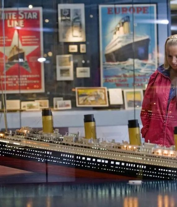 A girl in a pink jacket looks at a detailed model of the Titanic in a museum, with vintage posters and artefacts displayed on the walls behind glass.