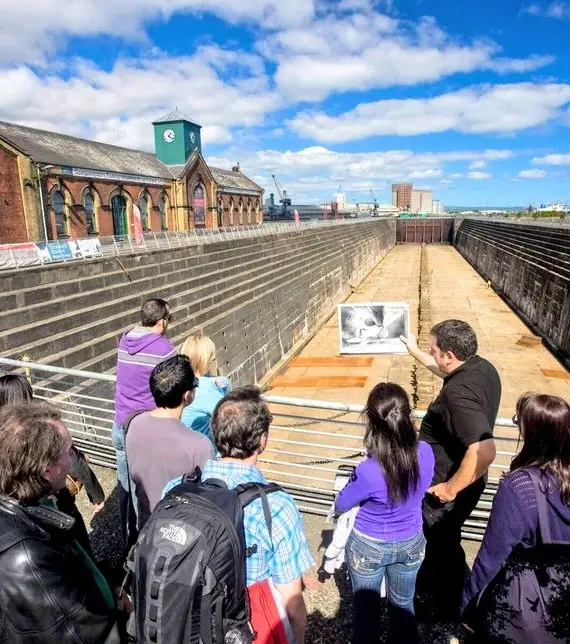 A group of people look into a large, empty dry dock surrounded by metal railings, with a tour guide explaining something whilst holding a photo; old brick buildings and cranes are in the background under a partly cloudy sky.