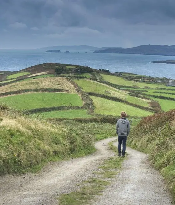 A person walks alone on a dirt path through green, patchwork fields, with rolling hills and the sea visible under a cloudy sky in the distance.