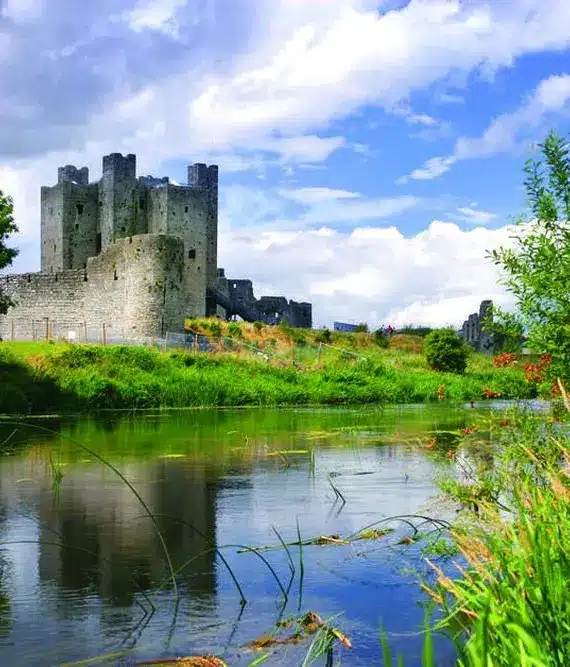 A stone castle with tall towers stands on a grassy hill beside a calm river, surrounded by lush green trees and plants under a partly cloudy blue sky.