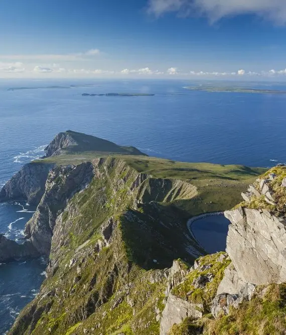 A person stands on a grassy cliff edge overlooking a dramatic coastal landscape with steep cliffs, rocky shores, and the vast blue sea under a partly cloudy sky.
