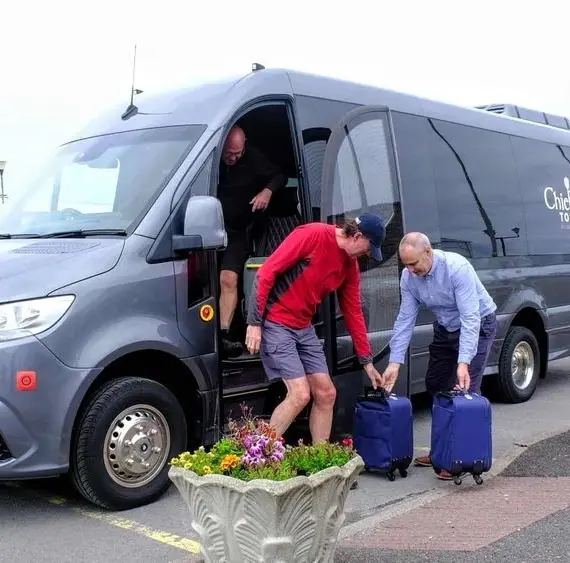 Three people unload blue suitcases from a dark grey tour van parked on a paved car park. Two are outside, one in a red jacket, and one is stepping out of the van. Flower planters are in the foreground.